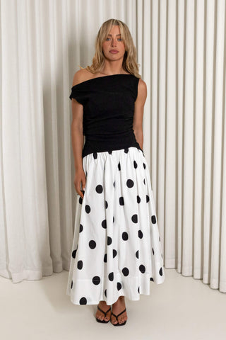 A woman modeling a black off-the-shoulder top paired with a white midi skirt featuring large black polka dots, standing in front of a white curtain backdrop.