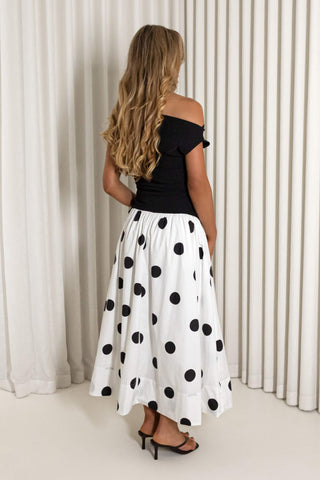 A woman shown from the back wearing a black off-the-shoulder top and a white midi skirt with large black polka dots, standing in front of white pleated curtains.