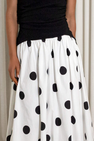 A white skirt with large black polka dots, worn by a model with a black top, against a neutral curtain background.
