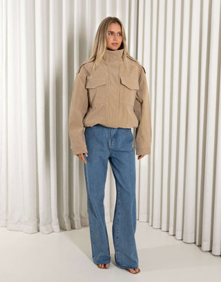 Woman wearing a beige jacket and blue jeans against a white curtain background