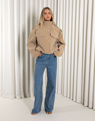Person wearing a beige jacket and blue jeans standing against a white curtain background