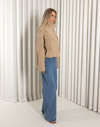Woman wearing a beige jacket and blue jeans standing against a white curtain.