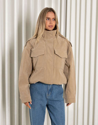 Person wearing a beige bomber jacket and blue jeans standing in front of white curtains