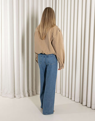 Person wearing a beige bomber jacket and blue jeans standing in front of white curtains