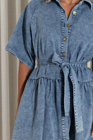 A close-up of a person wearing a blue denim shirt dress with short sleeves, a button front, and a tie waist belt.