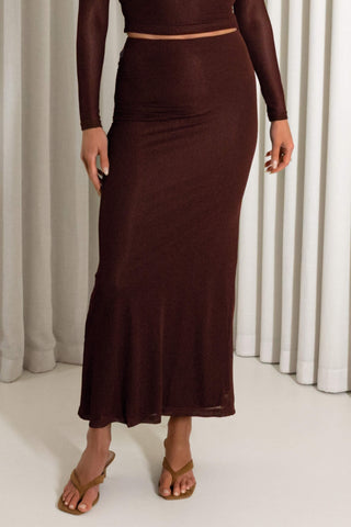 A woman wearing a dark brown long-sleeve midi skirt with a matching crop top, standing in front of white curtains.
