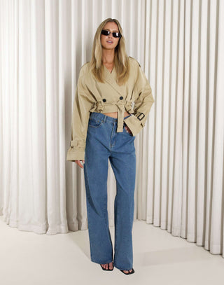 Woman wearing a beige cropped trench and denim jeans against a white curtain background