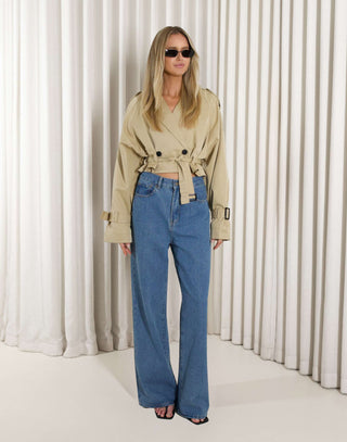 Woman wearing a beige cropped trench and denim jeans against a white curtain background