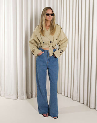 Woman wearing a beige cropped trench and denim jeans against a white curtain background