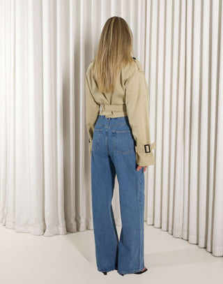 Woman wearing a beige cropped trench and denim jeans against a white curtain background