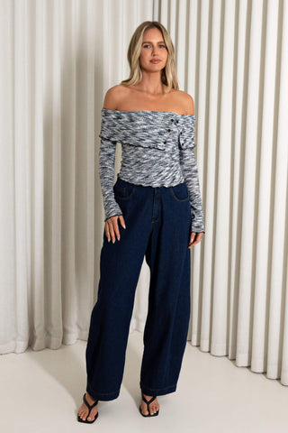 A woman modeling an off-the-shoulder long-sleeve top with a gray and white marled pattern, paired with dark blue wide-leg jeans and black sandals, standing in front of a cream-colored pleated curtain backdrop.
