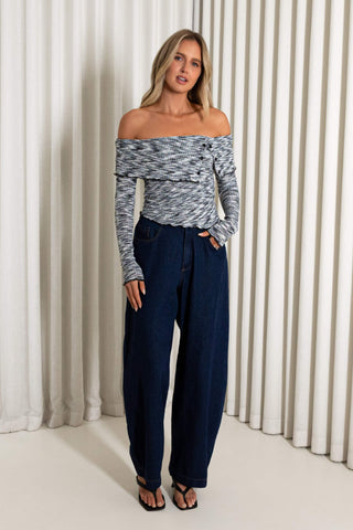 A woman modeling an off-the-shoulder long-sleeve knit top with a gray and white marled pattern, paired with dark blue wide-leg jeans and black sandals, standing in front of a cream-colored pleated curtain backdrop.