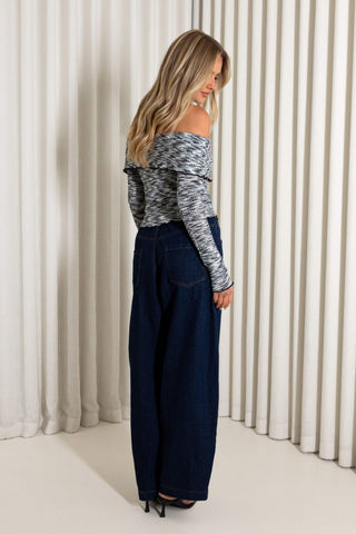 A woman is shown from the back wearing a gray and white patterned off-the-shoulder long-sleeve top and dark blue wide-leg denim jeans, standing in front of a white pleated curtain backdrop.