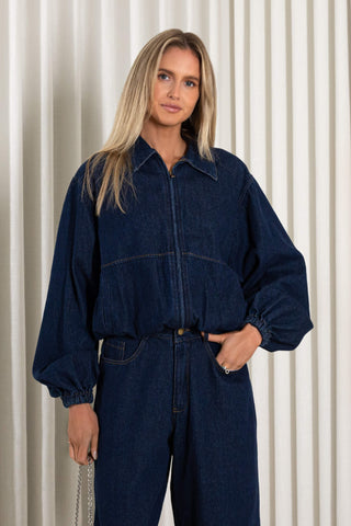 A woman modeling a dark indigo denim jumpsuit with a front zipper, collar, and voluminous long sleeves, standing against a cream-colored vertically pleated backdrop.