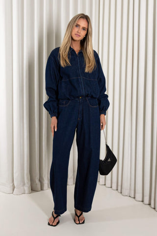 A woman modeling a dark indigo blue denim jumpsuit with front zip and button details, standing in front of a white pleated curtain backdrop.