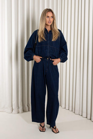 A woman modeling a dark navy blue denim jumpsuit with a front zipper, wide sleeves, and wide-leg pants, standing against a white pleated curtain backdrop.