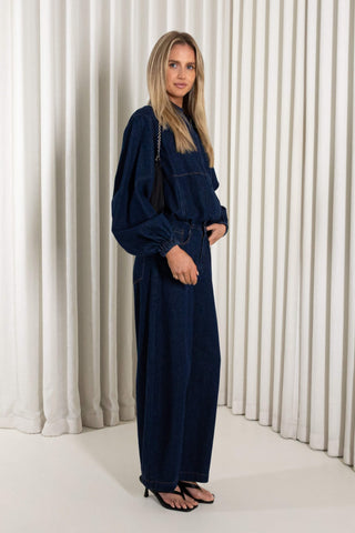 A woman modeling a dark navy blue denim jumpsuit with a front zipper, wide sleeves, and wide-leg pants, standing against a white pleated curtain backdrop.