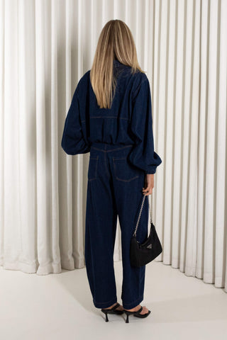 A woman modeling a dark navy blue denim jumpsuit with a front zipper, wide sleeves, and wide-leg pants, standing against a white pleated curtain backdrop.