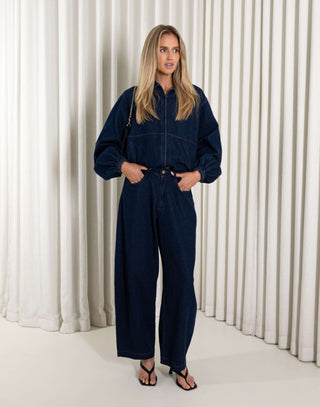 Woman wearing a denim blue jacket and matching pants standing against a white curtain background