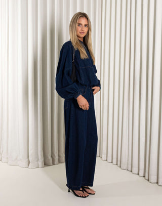 Woman wearing a denim blue jacket and matching pants standing against a white curtain background
