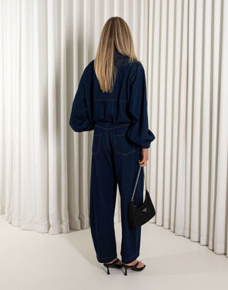 Woman wearing a denim blue jacket and matching pants standing against a white curtain background