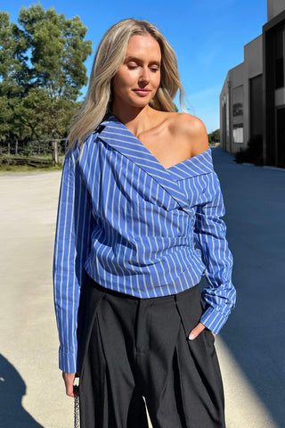 A woman wearing the blue and white vertically striped off-the-shoulder Perez long sleeve shirt style, paired with black wide-leg trousers, standing outdoors on a sunny day.