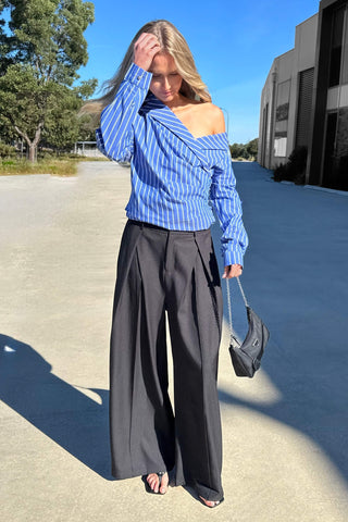 A woman wearing the blue and white vertically striped off-the-shoulder Perez long sleeve shirt style, paired with black wide-leg trousers, standing outdoors on a sunny day.