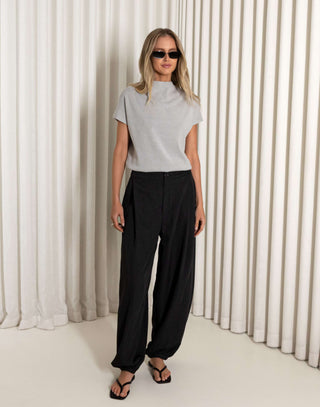 Woman wearing a gray top and black pants standing against a white curtain.