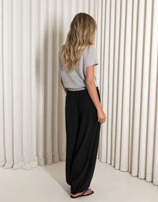 Woman wearing a gray top and black pants standing against a white curtain.