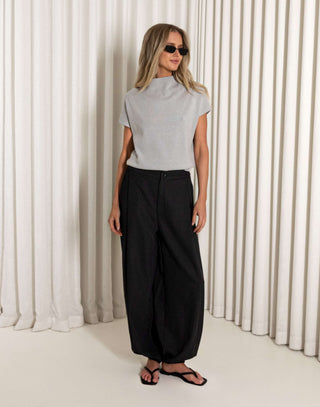 Woman wearing a gray top and black pants standing against a white curtain.