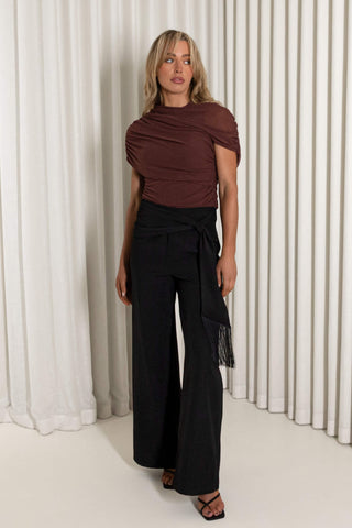 A woman modeling a dark brown draped short-sleeve top and black wide-leg trousers with a fringed tie detail, standing in front of white pleated curtains.
