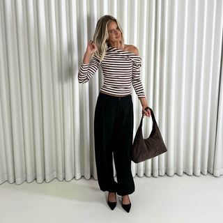 Woman wearing a brown and white striped top with black pants, holding a brown bag against a white curtain background.