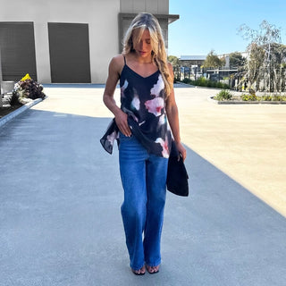 A woman stands outdoors on a paved walkway wearing a black spaghetti-strap camisole top with a pink, white, and gray floral print, paired with wide-leg blue jeans and holding a black clutch.
