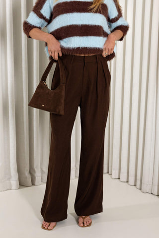 A person wearing high-waisted, wide-leg brown pants, a brown and light blue striped fuzzy sweater, and carrying a brown suede shoulder bag, standing in front of white curtains.