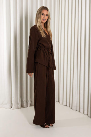 A woman modeling a dark brown belted wrap jacket and matching wide-leg trousers, standing against a white pleated curtain backdrop.