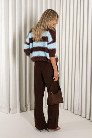 A woman stands with her back to the camera wearing a brown and light blue striped fuzzy cropped sweater, dark brown wide-leg trousers, and holding a matching dark brown handbag against a white pleated curtain backdrop.