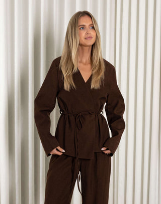 Woman wearing a brown blazer and matching pants standing against a white curtain background