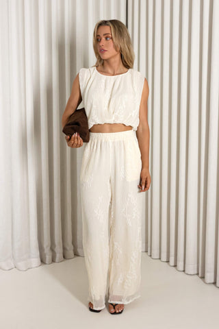 A woman modeling a two-piece cream lace outfit consisting of a sleeveless crop top and matching wide-leg pants, standing in front of a white pleated curtain backdrop.
