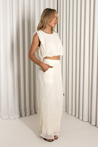A woman modeling a two-piece white outfit consisting of a sleeveless cropped top and matching high-waisted maxi skirt, standing in front of a white pleated curtain backdrop.