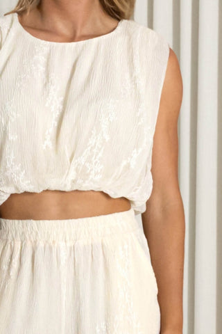 A close-up of a model wearing a sleeveless cream-colored textured crop top and matching high-waisted pants against a neutral curtain background.