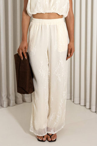 A model wearing a pair of off-white lace wide-leg pants with a matching crop top, holding a brown clutch bag in front of a neutral curtain backdrop.