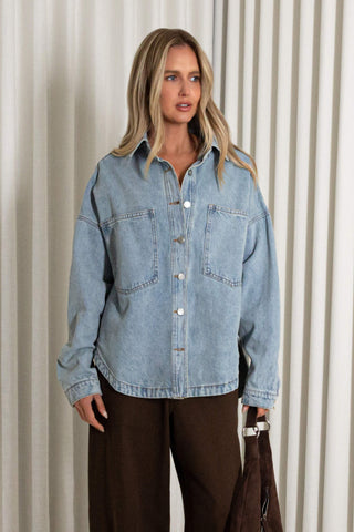 A woman wearing an oversized light blue denim button-up shirt with front chest pockets, paired with dark brown pants, standing against a beige pleated curtain backdrop.