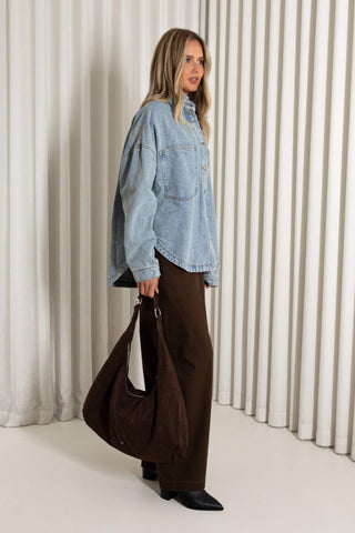 A woman stands in a studio wearing an oversized light-wash denim jacket, dark brown wide-leg pants, and black pointed shoes, while carrying a large dark brown slouchy shoulder bag. The background features vertical cream-colored drapes.