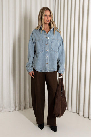 A woman modeling an oversized light blue denim shirt jacket with front buttons, paired with dark brown wide-leg trousers and holding a brown tote bag, set against a background of cream pleated curtains.