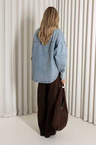 A woman viewed from behind wearing an oversized light blue denim shirt and dark brown wide-leg pants, holding a brown tote bag against a background of vertical cream-colored drapes.
