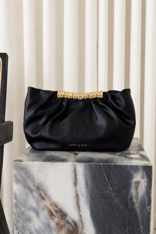 A black satin shoulder bag with a gold-tone beaded top handle and a small gold 'LUSTRE & SAGE' logo, displayed on a white and gray marble block against a cream pleated curtain background.