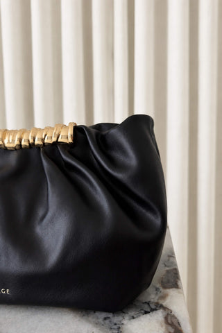 A close-up of a black leather shoulder bag with a distinctive gold metallic ring handle, resting on a marble surface against a background of vertical cream drapery.