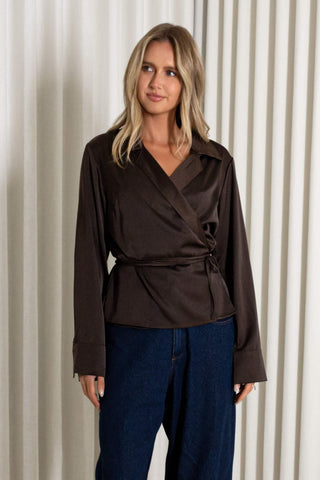 A woman modeling a dark brown satin wrap blouse with a tie waist, paired with dark blue jeans, standing against a cream-colored pleated curtain backdrop.