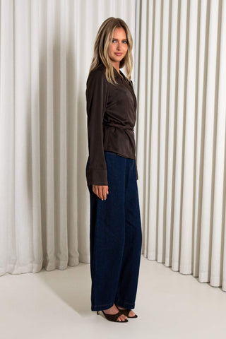 A woman modeling a dark brown long-sleeve wrap-style top and navy wide-leg pants against a background of vertical white drapes.