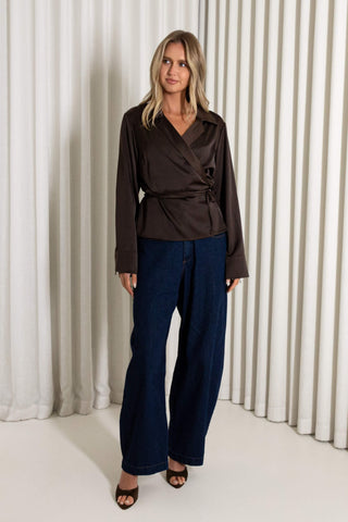 A woman modeling a dark brown satin wrap-style blouse with long sleeves and dark blue wide-leg trousers, standing in front of a white pleated curtain backdrop.